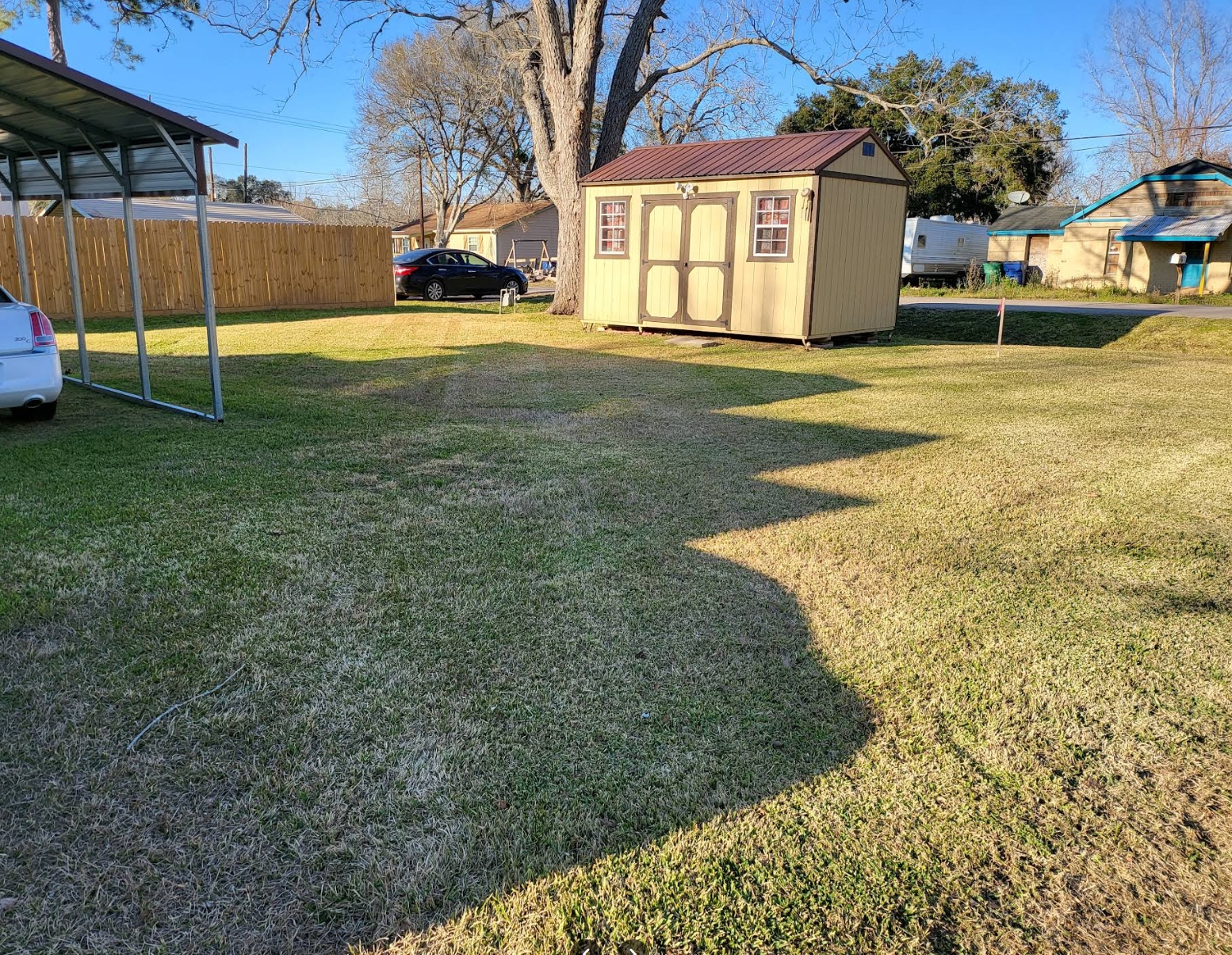 A back yard of dormant winter grass cut down evenly, with a cream storage shed under a red metal roof, an open metal carport and a wood privacy fence