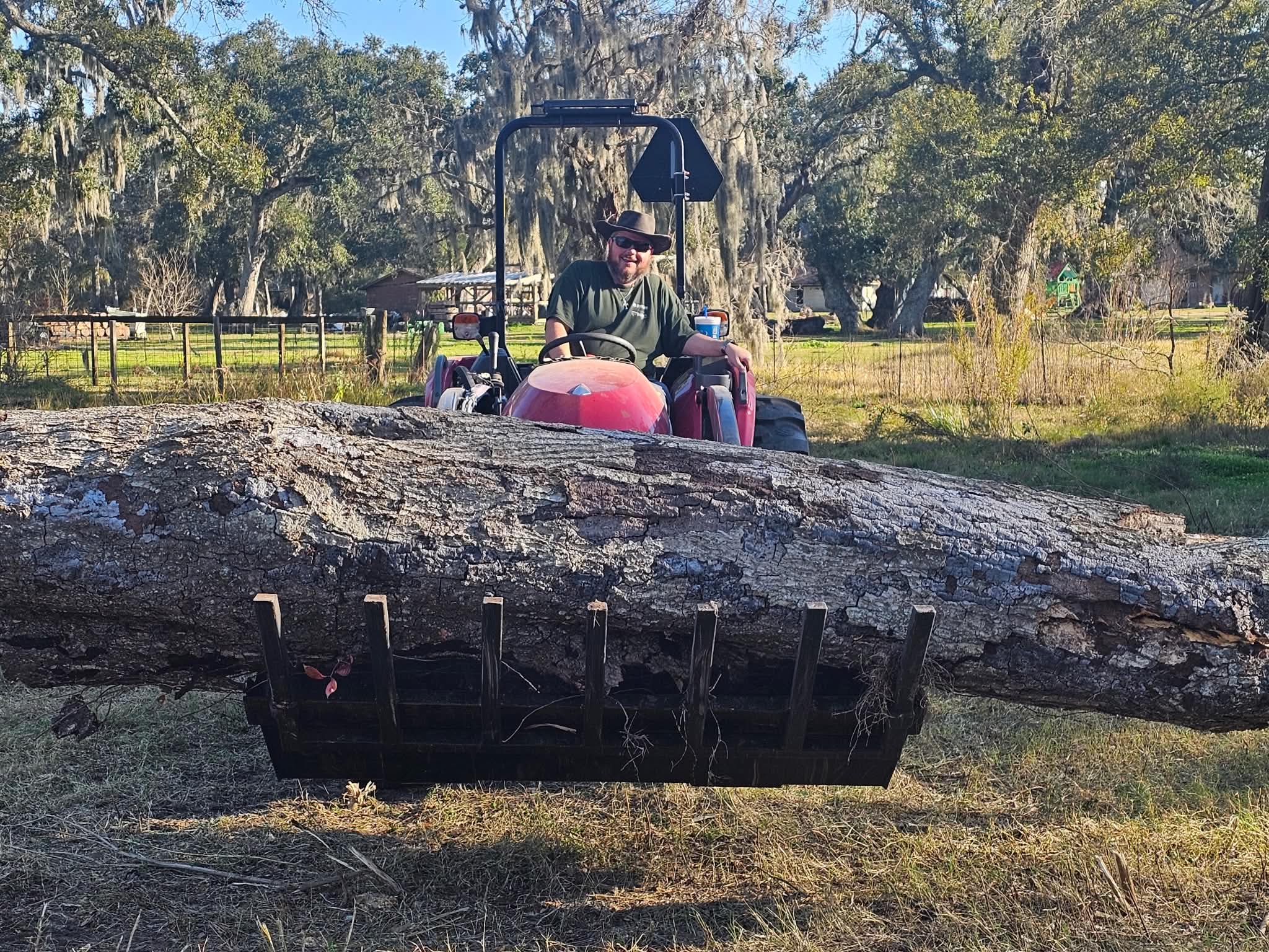 Mark on the red Yanmar tractor, smiling at the camera, carrying a long log across the pallet forks