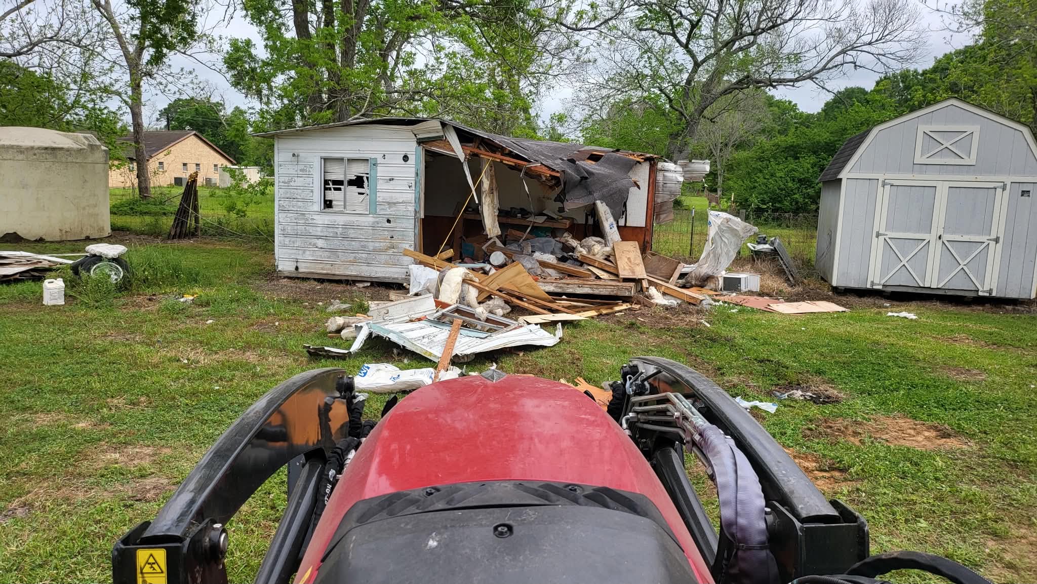 Seen from the tractor seat: a collapsed white clapboard outbuilding with timber, window frames and insulation spread across the grass