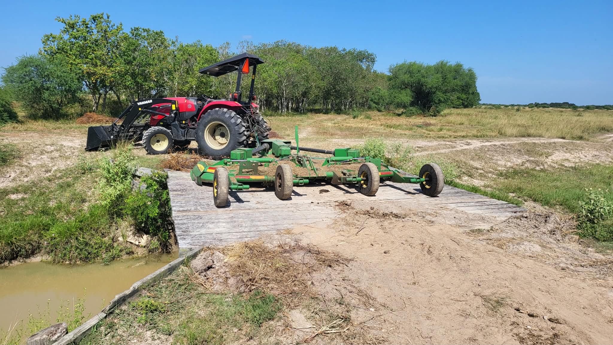 The red Yanmar tractor with a green batwing mower folded up behind it, parked on a timber bridge over a ditch on open pasture